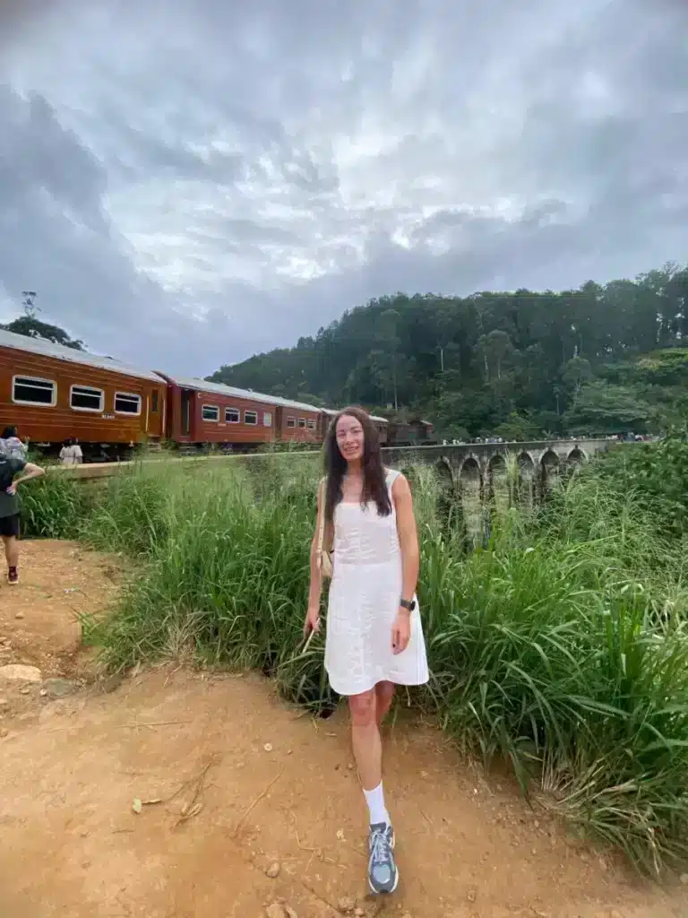 A woman in a white dress standing on a dirt path beside a passing train of reddish-brown carriages, with a stone viaduct and wooded hills behind