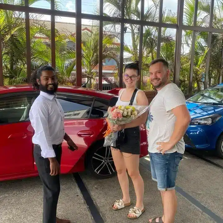 A man in a white shirt standing with a couple, the woman holding a bouquet, beside a red car under a canopy with palm trees behind