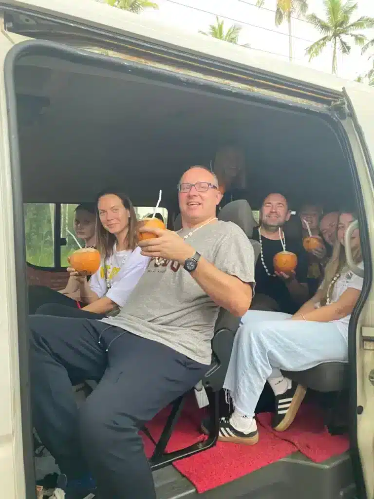 A group of travellers sitting in the open side door of a van, several of them holding orange king coconuts with straws