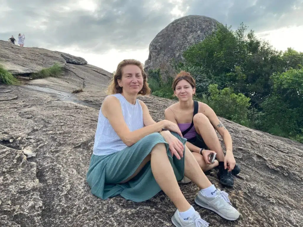 Two women sitting on a large granite rock, with a boulder and green shrubs behind them under a grey, cloudy sky