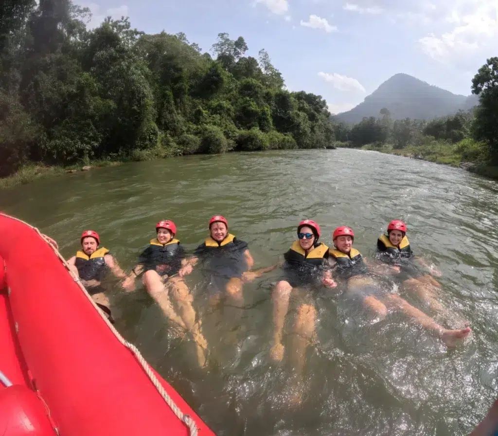 Six people in red helmets and yellow and black life jackets floating in a wide, calm river beside a red raft, with forest and a pointed hill behind