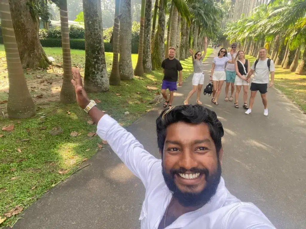A smiling man in white taking a selfie with a group of travellers posing on a palm-lined avenue