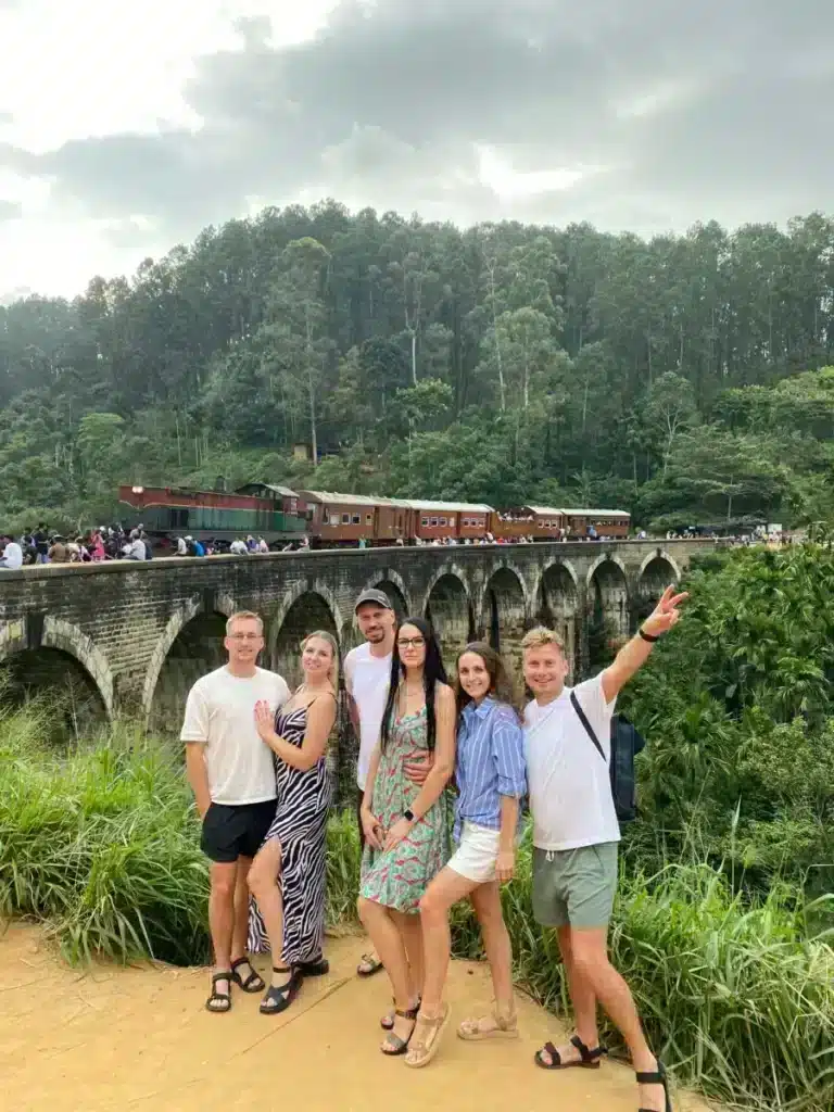 Six travellers posing on a dirt path in front of the Nine Arches Bridge while a train of reddish-brown carriages crosses it and people stand beside the track