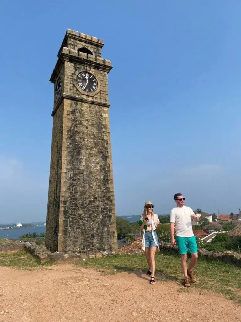 A man and a woman walking past an old stone clock tower on a grassy bank beside a low stone wall, with the sea on the left under a clear blue sky