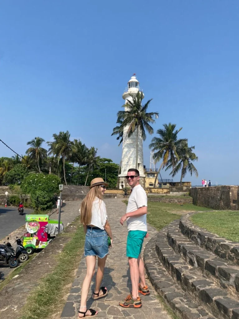 A couple on a paved path turning back towards the camera, with a white lighthouse and palm trees behind them under a cloudless sky