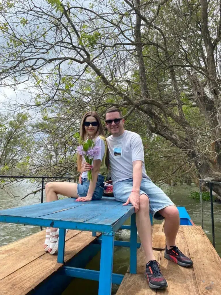 A couple sitting on a blue table on a wooden deck by the water, under a spreading tree, the woman holding purple flowers