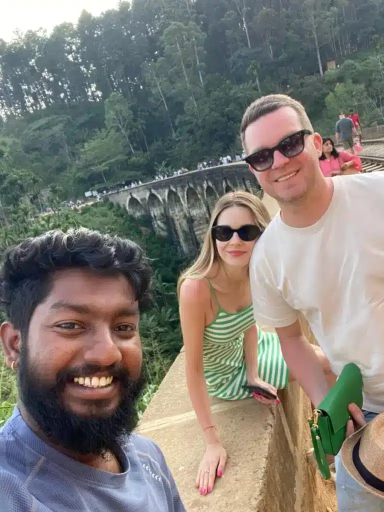 A smiling bearded man takes a selfie with a woman and a man in sunglasses at a low wall, with a long stone viaduct of arches and people walking along it among tall trees behind them