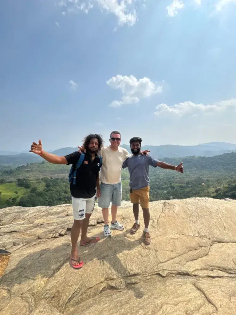 Three men arm in arm on a bare rock top, the two on the outside stretching an arm out, green hills behind them