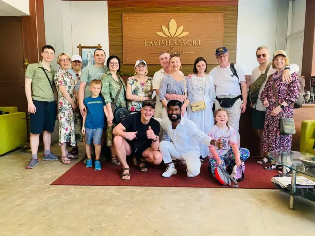 A large group of adults and children posing together on a red carpet in a hotel lobby, some crouching at the front and giving a thumbs up