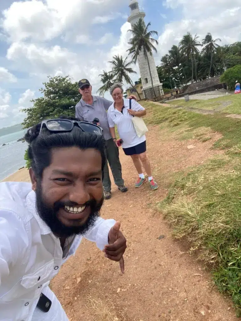A guide taking a selfie with two travellers on a grassy path, with a white lighthouse and the sea behind them
