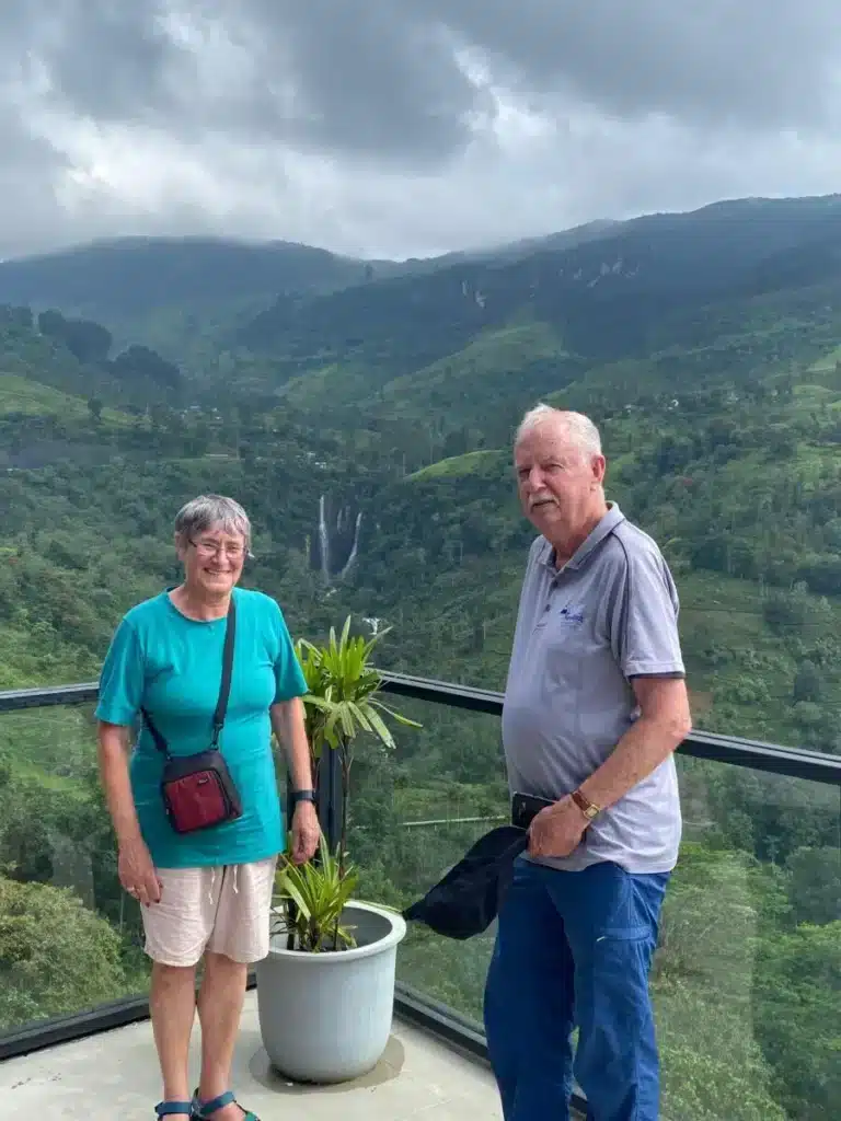 A man and a woman standing at a glass railing with a wide valley of green hills behind them, a waterfall in the distance and low grey cloud overhead