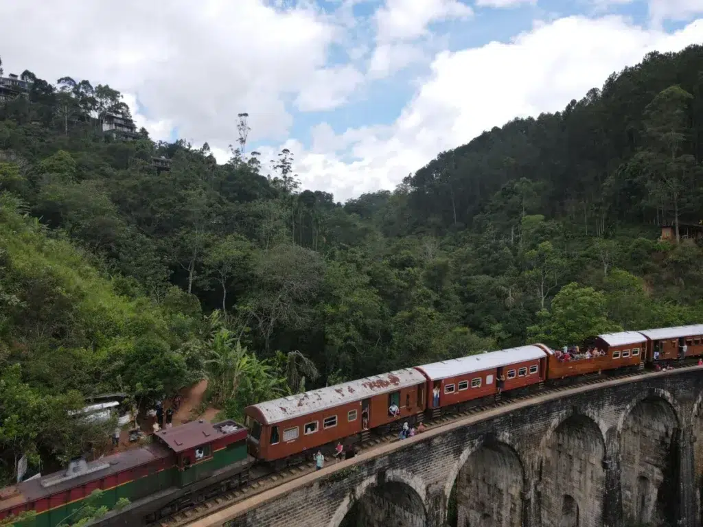 A train of reddish-brown carriages crossing a curved stone viaduct with tall arches, surrounded by forested hills