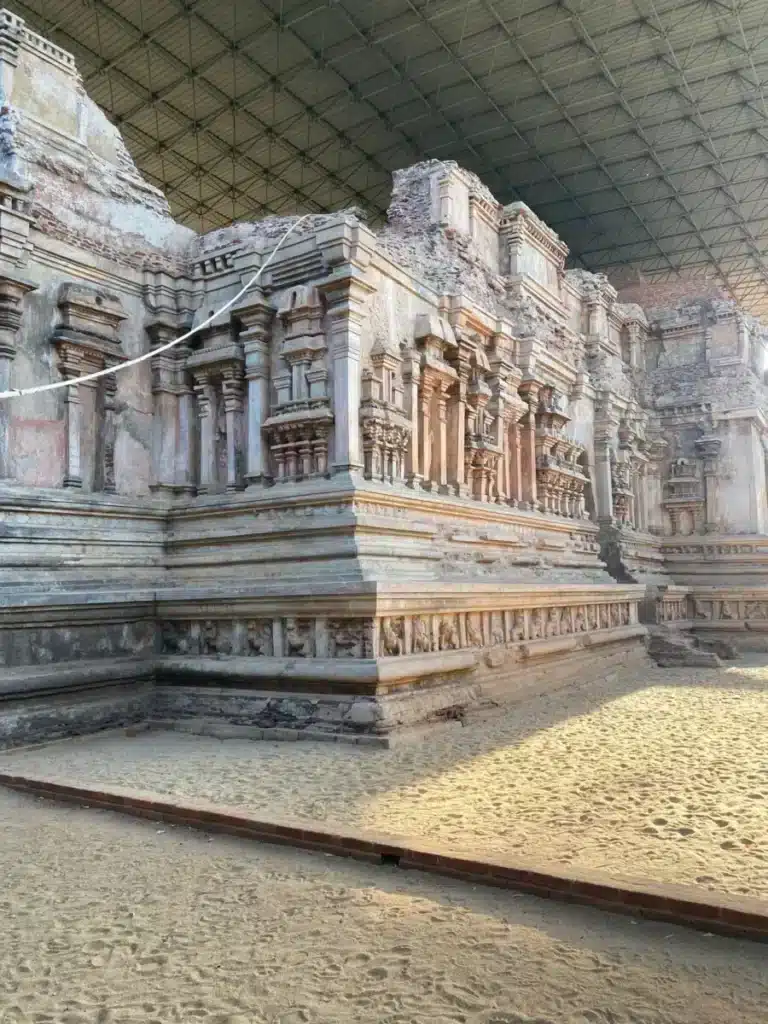 An ornate ruined shrine with rows of pilasters and a band of small figures along its base, standing on sand under a large steel roof