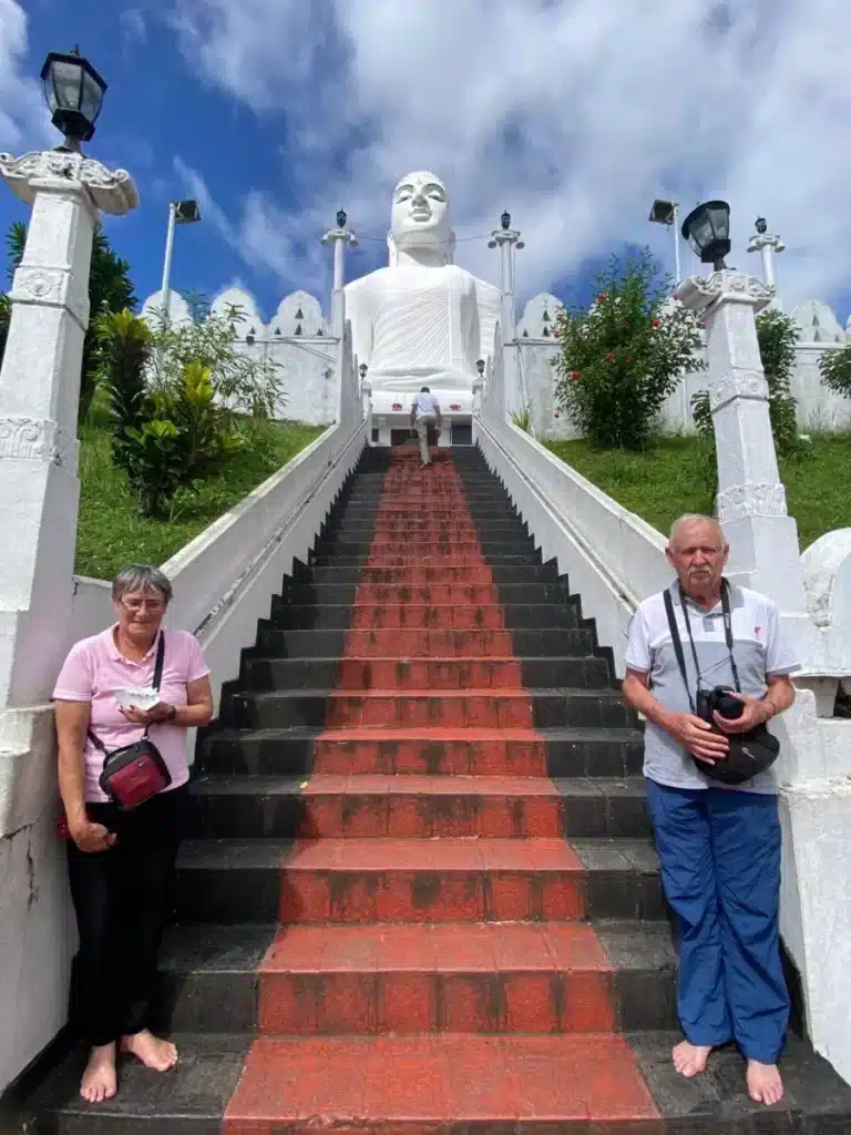 Two visitors standing barefoot on a staircase with a red strip down the middle, leading up to a large white seated Buddha statue