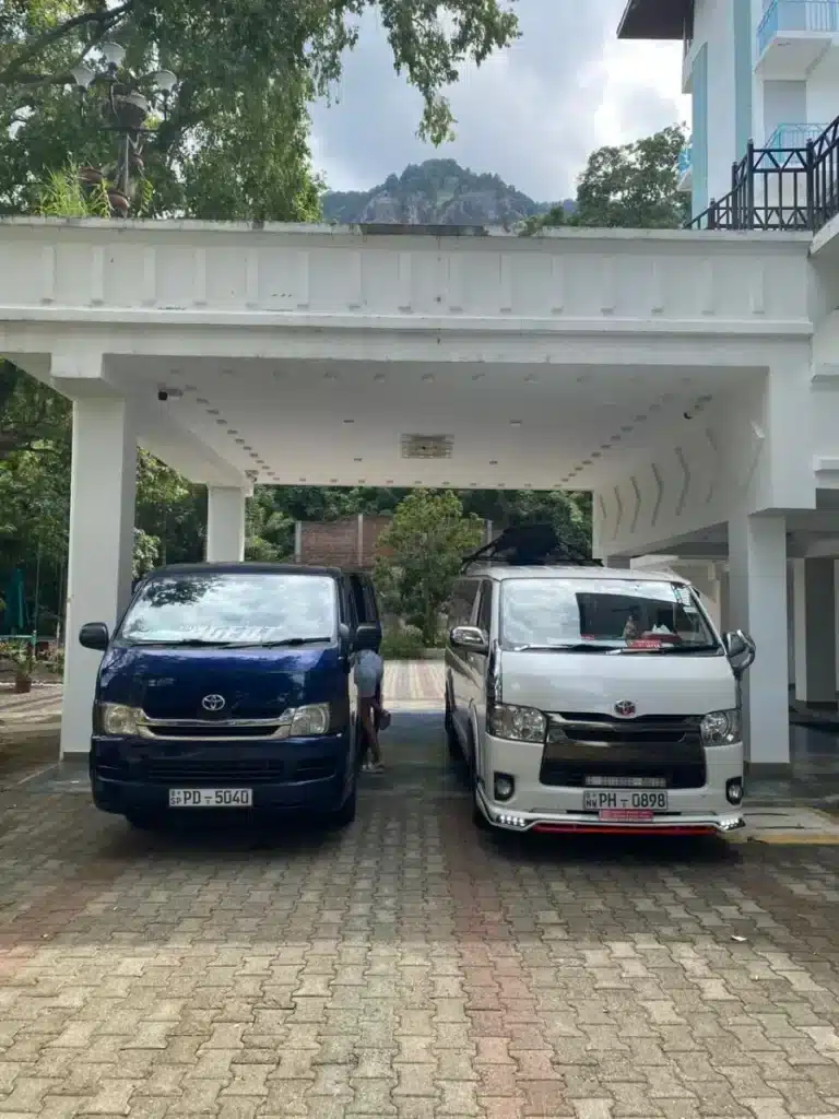 A dark blue van and a white van parked side by side under a white porch on a block-paved forecourt, with a wooded rocky hill behind