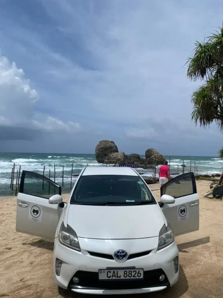 A white car with its doors open on a sandy beach, with a rocky islet and stilt-fishing poles in a rough sea behind it