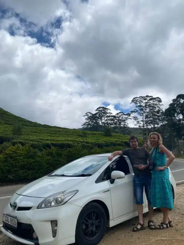 A man and a woman leaning on a white car parked beside a road, with a tea-covered slope and tall trees behind them under a cloudy sky